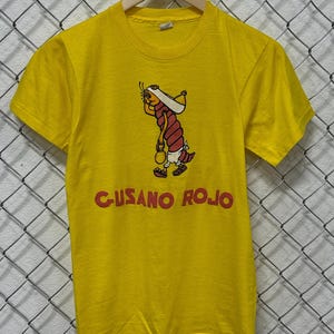 Könnte beinhalten: Gelbes T-Shirt mit einer Cartoon-Illustration eines roten, wurmartigen Charakters, der eine Tasche trägt und Sandalen trägt. Der Text "GUSANO ROJO" ist rot gedruckt. Das T-Shirt hängt an einem Holzbügel.