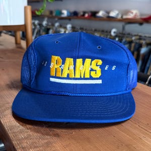May include: A royal blue trucker hat featuring a mesh back and a flat brim. The front panel displays the yellow embroidered text RAMS with the white text LOS ANGELES arched above it and a horizontal white line underneath the word RAMS.