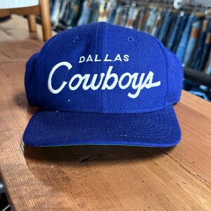 Op de afbeelding: Een koningsblauwe honkbalpet met de tekst Dallas Cowboys in het wit geborduurd op de voorkant. De pet heeft een gestructureerd ontwerp met zes panelen, een platte klep en een groene onderkant, wat een klassieke sportieve stijl uitstraalt.