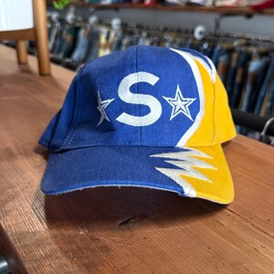 May include: A blue baseball cap featuring a large white embroidered letter S flanked by two white stars on the front panel. The right side of the cap is yellow with a white lightning bolt graphic extending from the brim up to the side panel.
