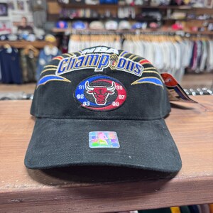 May include: A black baseball cap featuring an embroidered Chicago Bulls logo surrounded by the years 91, 92, 93, 96, 97, and 98. The text NBA Champions is embroidered in blue and gold above the logo, with blue and gold lightning bolt accents.