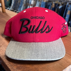Puede incluir: Una gorra de béisbol roja con visera plana gris. La parte frontal presenta el texto CHICAGO Bulls bordado en negro. Un pequeño logotipo de los Chicago Bulls en blanco está bordado en el panel lateral derecho. La corona tiene ojales blancos.