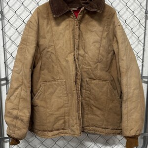 Puede incluir: Chaqueta acolchada de color canela con cuello de pana marrón y dos bolsillos delanteros. La chaqueta tiene un forro rojo y puños acanalados marrones. Cuelga de una percha de madera contra una valla de alambre.