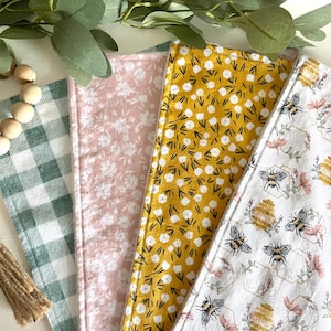 Könnte beinhalten: Vier Stoffquadrate mit verschiedenen Mustern. Ein Quadrat ist grün-weiß kariert, eines ist rosa mit weißem Blumenmuster, eines ist gelb mit schwarzem Blumenmuster und eines ist weiß mit einem Bienen- und Wabenmuster.