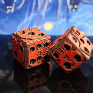 May include: Two red dice with black detailing and skull-like designs. The dice are made of a textured material and have a slightly worn appearance.