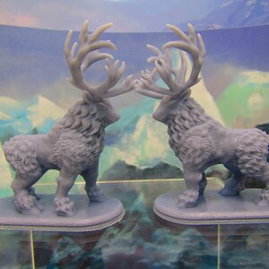May include: Two gray, furry, miniature figurines of deer with large antlers. The deer are facing each other and are standing on small, gray bases.