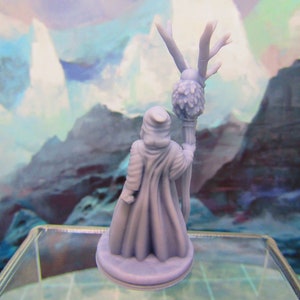 May include: A miniature figurine of a robed figure holding a staff with a leafy top. The figure is painted in a light blue color and is standing on a round base.