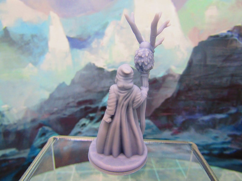 May include: A miniature figurine of a robed figure holding a staff with a leafy top. The figure is painted in a light blue color and is standing on a round base.