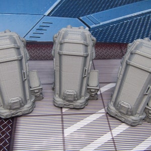 May include: Three gray 3D printed model coffins with a detailed design. The coffins are sitting on a gray and white striped surface.