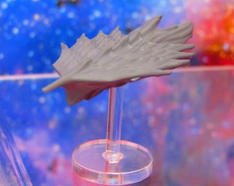 May include: A gray, wing-shaped sculpture mounted on a clear acrylic stand. The sculpture has a textured surface with detailed ridges and a pointed tip. The stand has a circular base and a clear, cylindrical support.