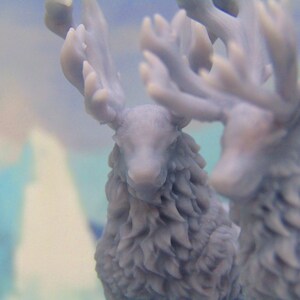 May include: Two gray, furry reindeer figurines with antlers. The reindeer are facing forward with their heads tilted slightly down. The background is a blurry blue and white.