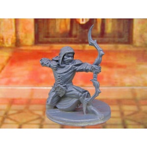 May include: A gray miniature figurine of an archer in a hooded cloak, kneeling and drawing a bow. The archer is on a round base. The bow has a decorative, curved design. The figurine is set against a blurred background.