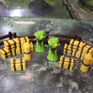May include: A miniature farm scene with a brown wooden fence, yellow hay bales, sheaves of wheat, and bundles of wheat. The scene is set on a green and gray surface, creating a rustic, countryside theme.