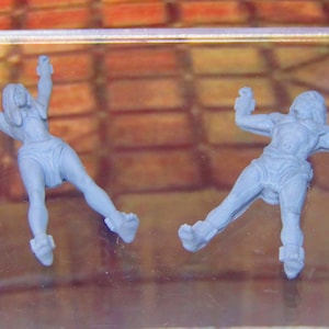 May include: Two gray plastic figurines of people with their arms bound behind their backs. The figures are lying on a glass surface.