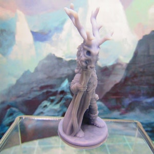 May include: A gray, 3D printed miniature figurine of a creature with a deer skull mask and antlers, wearing a cloak and holding a staff. The figurine is standing on a round base.