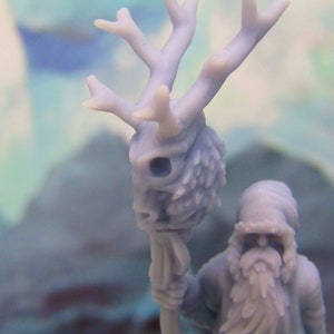 May include: A miniature figurine of a bearded figure holding a staff topped with a deer skull. The skull is detailed with antlers and a textured surface.