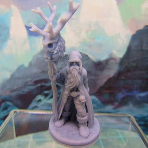 May include: A miniature figurine of a bearded figure wearing a hooded cloak and holding a staff topped with a deer skull. The figure is painted in a light blue color and stands on a round base.