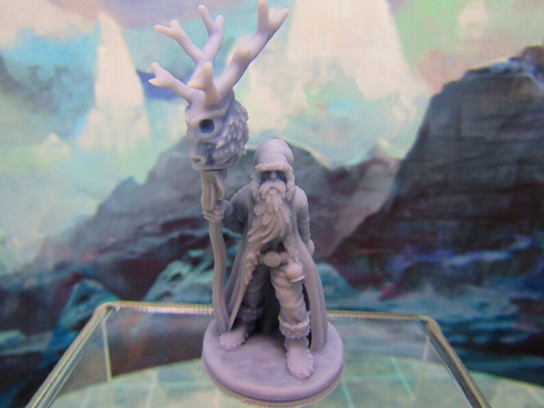 May include: A miniature figurine of a bearded figure wearing a hooded cloak and holding a staff topped with a deer skull. The figure is painted in a light blue color and stands on a round base.