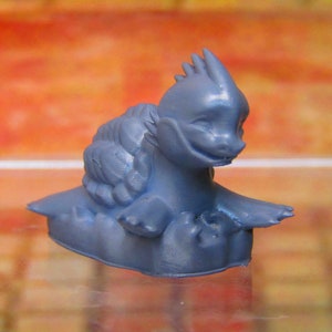 May include: A blue plastic figurine of a small, spiky, dragon-like creature with a wide mouth and a friendly expression. The creature is sitting on a small, rounded base.