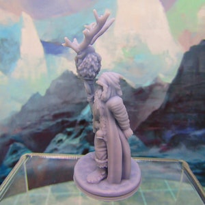 May include: A miniature figurine of a person wearing a hooded cloak and holding a staff with a deer head on top. The figurine is made of a light gray plastic and is standing on a round base.