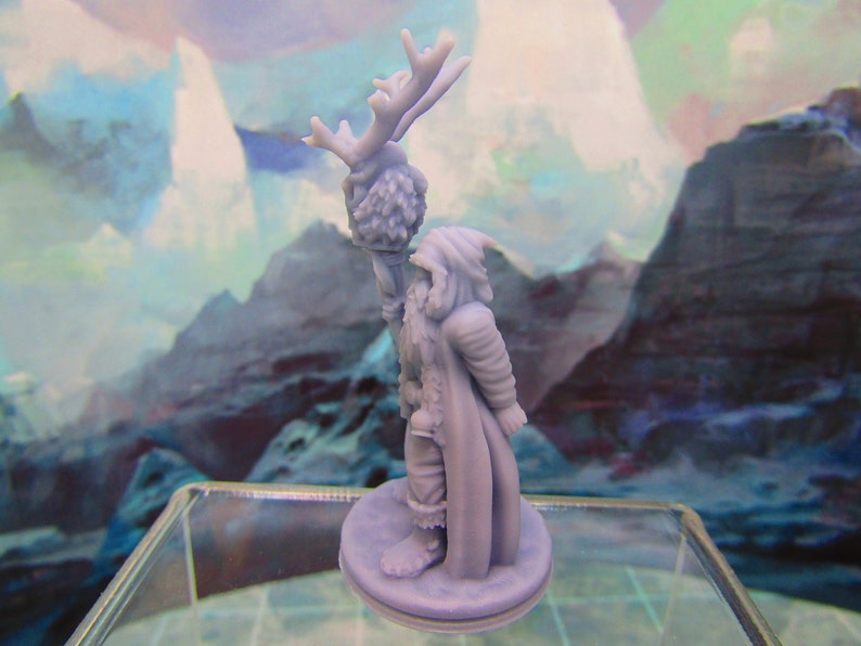 May include: A miniature figurine of a person wearing a hooded cloak and holding a staff with a deer head on top. The figurine is made of a light gray plastic and is standing on a round base.