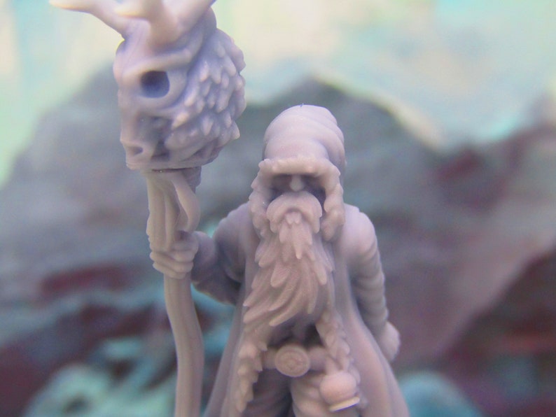 May include: A miniature figurine of a bearded figure wearing a hooded cloak and holding a staff topped with a skull. The figure is painted in a light purple color.