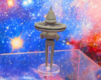 May include: A gray space station model with a central hub and a ring around it, on a clear plastic stand against a blue and red starry background.