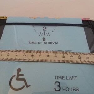 May include: A blue parking permit with a wheelchair symbol, a time of arrival indicator, and a time limit of 3 hours. The permit is displayed on a brown ruler.