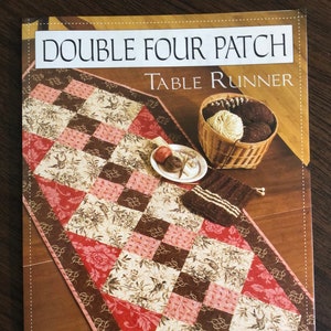 Pode incluir: Um padrão de acolchoado para um corredor de mesa chamado "Double Four Patch Table Runner" da Graywood Designs. O padrão apresenta um tecido floral marrom, rosa e creme com uma borda marrom e rosa.