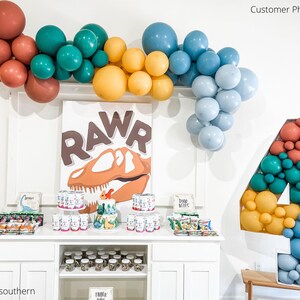 May include: A dinosaur-themed party setup with a balloon arch in shades of red, teal, yellow, and blue. A large number "4" is filled with balloons. A sign says "RAWR" with a dinosaur graphic. Party supplies are on display.