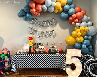 May include: A race car themed party decoration set with a checkered tablecloth, a large number 5 light-up sign, a cake, and a balloon arch in blue, yellow, and orange. The balloons spell out "Welcome".
