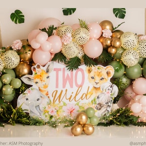 May include: A pink and green balloon arch with a sign that says "Two Wild" in pink letters. The arch is decorated with animal illustrations, including a giraffe, lion, and zebra.