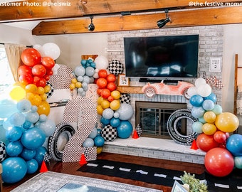 May include: A race car themed party decoration with a large checkered number two, a red toy car, and a balloon arch in blue, yellow, red, and white. The balloon arch is decorated with checkered flags and tires.