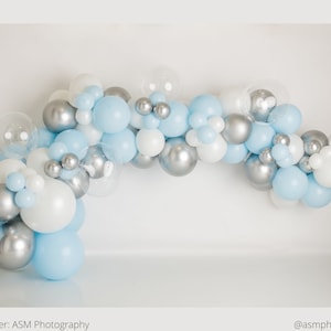 May include: A balloon arch made of silver, white, and light blue balloons. The arch is curved and hangs from a white wall.