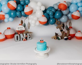 Puede incluir: Una escena festiva de primer cumpleaños con una tarta azul coronada con un flotador de pesca. El fondo incluye un arco de globos en tonos azules y blancos, flotadores de pesca decorativos y un ancla. Se muestra la palabra "ONE".