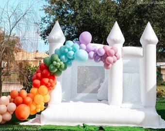 Op de afbeelding: Witte opblaasbare springkussen met een regenboogballonboog. Het springkussen heeft een wit gaasvenster en twee witte torens. De ballonboog is gemaakt van rode, oranje, gele, groene, blauwe, paarse en roze ballonnen.