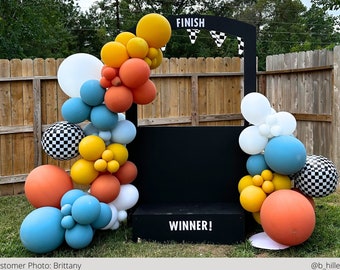 May include: A black and white checkered finish line backdrop with a black sign that says "FINISH" and "WINNER!" decorated with a balloon arch of orange, yellow, blue, and white balloons.