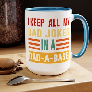 May include: White ceramic mug with a blue rim and handle. The mug has a retro-style design with the text "I keep all my dad jokes in a dad-a-base".