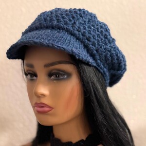 May include: A blue crocheted newsboy cap with a textured design. The cap features a small brim and a rounded crown. The hat is made of a soft, textured yarn. The cap is shown on a mannequin with black hair and a black choker.