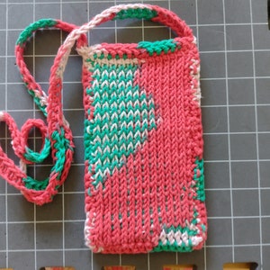 May include: A knitted phone case with a pink, green, and white color scheme. The case has a long strap for carrying it around the neck.