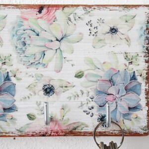 May include: A white wooden key holder with a floral design featuring blue and pink succulents and white flowers. The key holder has five silver hooks.