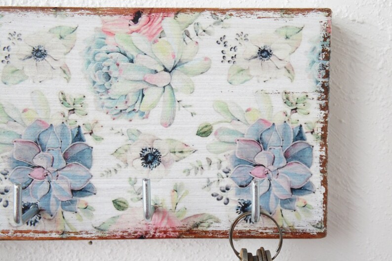 May include: A white wooden key holder with a floral design featuring blue and pink succulents and white flowers. The key holder has five silver hooks.