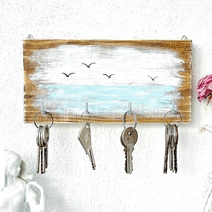 May include: A rustic wooden key holder with a painted beach scene. The design features a white sky, a light blue ocean, and black bird silhouettes. Four metal hooks hold sets of keys, offering a practical and decorative storage solution.