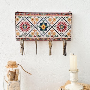 Może przedstawiać: Dekoracyjny wieszak na klucze z kolorowym wzorem płytek. Prostokątny uchwyt ma powtarzający się wzór czerwonych, żółtych i niebieskich motywów kwiatowych. Poniżej przymocowano pięć metalowych haczyków, każdy z zestawem kluczy. Uchwyt jest zamontowany na białej ścianie.