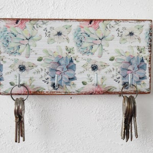 May include: A wooden key holder with a floral design featuring succulents and flowers in shades of pink, blue, and green. The holder has three hooks for hanging keys.