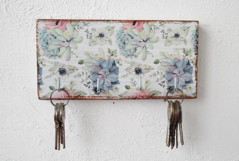 May include: A wooden key holder with a floral design featuring succulents and flowers in shades of pink, blue, and green. The holder has three hooks for hanging keys.