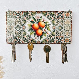 May include: A decorative key holder featuring a vibrant orange and floral design. The rectangular wooden board has a tile-like pattern with a central illustration of oranges and blossoms. Four metal hooks hold keys, adding functionality to the artwork.