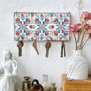 Puede incluir: Un porta llaves de madera blanco con un patrón floral colorido. El soporte tiene cinco ganchos para colgar llaves. Hay cinco llaves colgadas de los ganchos.