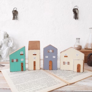 May include: Four miniature wooden houses painted in different colours. The houses are arranged in a row on a white surface. The houses are all different sizes and shapes, and they have different details, such as windows, doors, and chimneys.