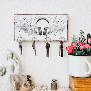 Puede incluir: Porta llaves de madera blanca con una ilustración en blanco y negro de auriculares y notas musicales. Cinco llaves cuelgan de ganchos en el soporte.